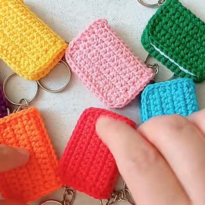 Puede incluir: Una colección de llaveros de ganchillo coloridos en varios colores, incluyendo amarillo, rosa, verde, azul, naranja, rojo y morado. Cada llavero es rectangular y está unido a un llavero plateado.