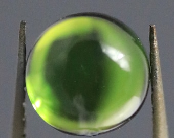 Puede incluir: Una gema redonda y translúcida con un degradado de tonos verdes y amarillos. La gema está sujeta por unas pinzas metálicas, que resaltan su superficie pulida y su forma esférica. El color pasa de un verde vibrante a un amarillo más claro.