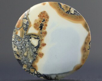 May include: A round, polished stone with a unique pattern. The stone features a large, light blue area, bordered by a mix of beige, brown, black, and gray speckled patterns. The overall effect is a natural, earthy aesthetic.