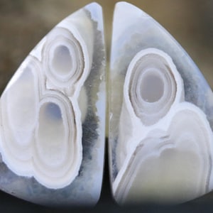May include: Two triangular cabochons of polished agate stone featuring a translucent light blue and white colour palette. The stones display distinct concentric banding patterns that resemble eye-like shapes with layered rings of white and grey.