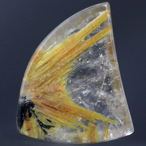 May include: A clear, polished gemstone with golden, needle-like inclusions radiating throughout. The stone has a rounded, triangular shape and a translucent quality, with a dark, black area at the base.