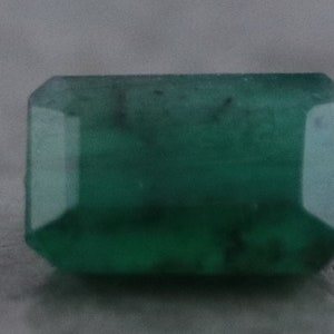 May include: A rectangular emerald-cut gemstone with a deep green hue. The gem has a faceted surface, reflecting light and showcasing its clarity. The stone's shape is a classic rectangular with cut corners.