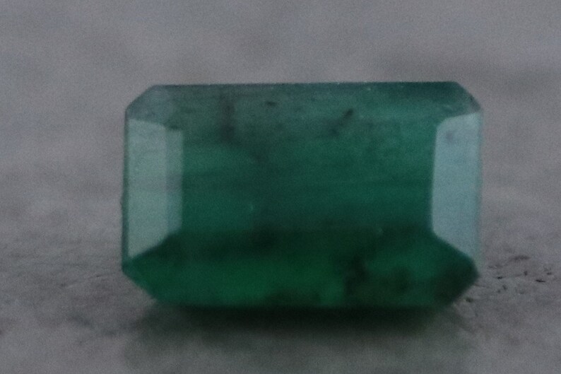 May include: A rectangular emerald-cut gemstone with a deep green hue. The gem has a faceted surface, reflecting light and showcasing its clarity. The stone's shape is a classic rectangular with cut corners.