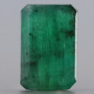 May include: A rectangular-cut emerald gemstone with a deep green hue. The gemstone has a faceted surface with visible inclusions, and a polished finish. The emerald is set against a neutral background, highlighting its color and clarity. This gemstone is suitable for jewelry making.