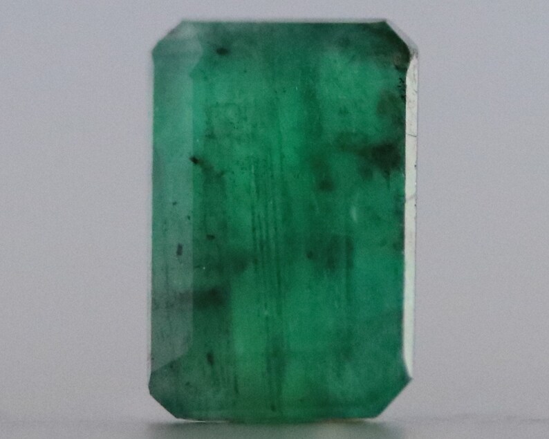 May include: A rectangular-cut emerald gemstone with a deep green hue. The gemstone has a faceted surface with visible inclusions, and a polished finish. The emerald is set against a neutral background, highlighting its color and clarity. This gemstone is suitable for jewelry making.