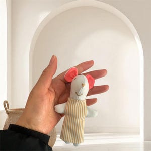 Puede incluir: Una muñeca de ratón blanca hecha a mano con orejas rosas y un corazón dorado. El ratón lleva un delantal a rayas en beige y blanco. La muñeca se sostiene en una mano, con una cesta tejida en el fondo.