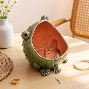 Puede incluir: Un cuenco de cerámica verde con forma de rana y un interior naranja. La rana tiene ojos grandes y piel texturizada. Varios anillos y un collar están dentro del cuenco. Un anillo de oro está sobre la mesa.