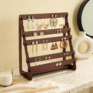 May include: A dark brown wooden earring display stand with multiple rows of holes, showcasing various earrings. The earrings include gold hoops, beaded dangles, and stud earrings. The stand is angled for visibility and sits on a light-colored surface.