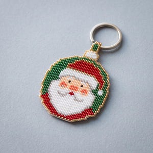 Puede incluir: Un llavero circular con un rostro de Santa Claus hecho de cuentas. El diseño utiliza cuentas rojas, blancas, verdes y color melocotón con un borde de cuentas doradas. Un anillo de metal plateado está sujeto a la parte superior del adorno.