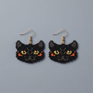 May include: A pair of dangle earrings featuring a beaded cat face design. The cat faces are primarily black with yellow eyes, orange cheek accents, and gray ear details. Each earring hangs from a gold-toned metal hook for pierced ears.
