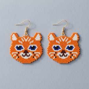 以下が含まれることがあります： ビーズで作られた猫の顔のデザインのドロップイヤリング。オレンジ、白、黒、青のビーズで構成されており、尖った耳、青い目、暗い鼻を持つ猫の顔が描かれています。各イヤリングはゴールドトーンのフックから吊り下げられています。