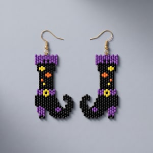 May include: A pair of beaded dangle earrings shaped like boots. The boots are primarily black with purple cuffs, heels, and toe tips. They feature small yellow and orange square accents on the shaft and a yellow buckle detail at the ankle.