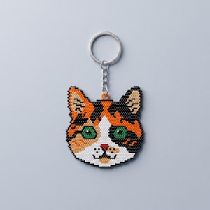 May include: A calico cat keychain crafted from small, colorful beads. The cat face features black, orange, and white fur patterns, bright green eyes, and a small red nose. A silver-toned metal split ring is attached to the top by a short chain.