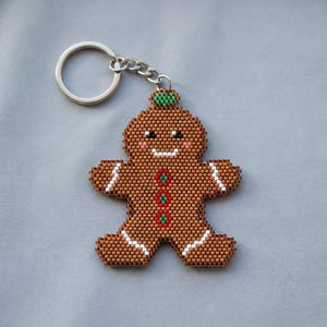 May include: A gingerbread person keychain crafted from brown, white, red, and green beads. The figure features a small green hat, pink cheeks, a white smile, and three red and green buttons on the chest, attached to a silver metal ring.