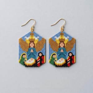 May include: A pair of hexagonal drop earrings featuring a beaded nativity scene. The design depicts a central figure in blue, a yellow star, and smaller figures in green and red, all crafted from tiny colourful beads with gold-tone metal hooks.