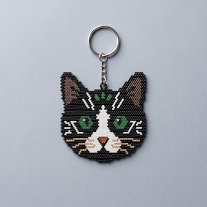 May include: A beaded keychain shaped like a cat head with black, white, and brown beads. The cat has green eyes, a small orange nose, and a white patch on its forehead and muzzle. A silver metal split ring is attached to the top by a short chain.