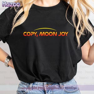 Może przedstawiać: Czarny t-shirt z napisem "COPY, MOON JOY" w gradientowej pomarańczowo-żółtej czcionce. Tekst jest łukowaty z żółtą linią nad nim. Koszulka ma okrągły dekolt.