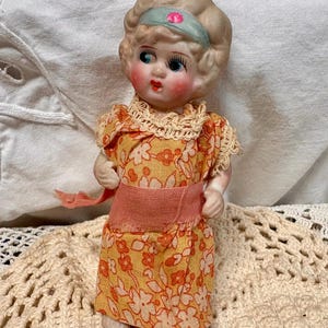 May include: A small porcelain doll with blonde hair, blue eyes, and rosy cheeks, wearing a light blue headband with a pink flower. The doll is dressed in a yellow and orange floral print dress with a peach-colored sash and cream lace trim.