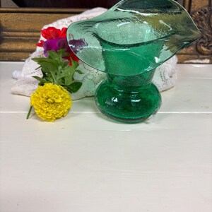 May include: A green glass vase with a unique, flared top. The vase has a textured surface and a rounded base. A yellow flower and other colorful flowers are next to the vase. The vase is on a white surface.