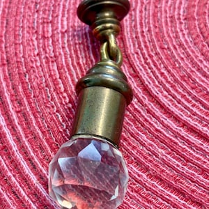 May include: A decorative bottle stopper with a faceted crystal ball base and antique brass hardware. The stopper features a detailed design with a cylindrical body and a decorative top. The crystal ball is clear and reflects light.