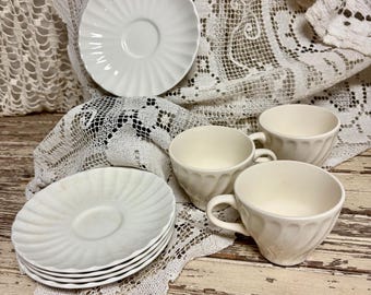 May include: A set of off-white ceramic tableware consisting of three fluted saucers and two matching cups with curved handles. The saucers feature a radial ribbed pattern, while the cups have a subtle vertical swirl design on their exterior.