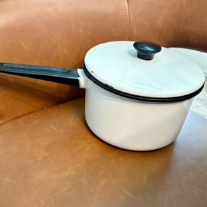 May include: A white enamel saucepan with a black rim and a matching lid featuring a black knob. The pan has a long, straight black handle extending from the side. The exterior finish is smooth and glossy, showing minor signs of wear along the edges.