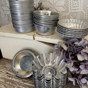 May include: A collection of vintage metal baking molds in various shapes and sizes. The molds include round, fluted, and ring-shaped designs, stacked on a white surface. The metal has a silver finish, and the arrangement suggests a focus on baking and kitchenware.