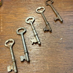 May include: Five antique metal keys of varying sizes and designs are arranged diagonally on a wooden surface. The keys have a tarnished, aged appearance, with intricate details on the key heads and teeth. The wood has a warm, brown tone.