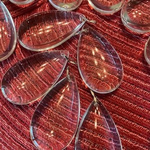 May include: Clear, teardrop-shaped glass ornaments with a textured, reddish-brown interior. Each ornament has a small metal loop at the top. The ornaments are arranged on a red, textured surface.