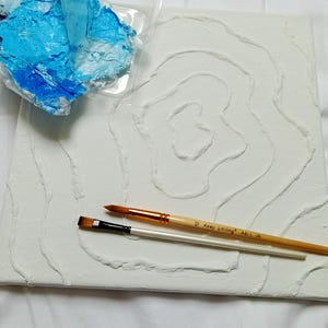 Könnte beinhalten: Eine weiße, strukturierte Leinwand mit einem erhabenen Blumendesign, zusammen mit zwei Pinseln und einem Plastikbehälter mit blauer Farbe. Die Pinsel haben Holzgriffe und liegen diagonal auf der Leinwand.