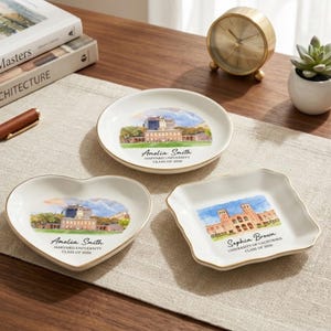 May include: Three white ceramic dishes with gold rims, each featuring a watercolor image of a university building. One is round, one heart-shaped, and one square. Each dish has a name and graduation year printed on it.