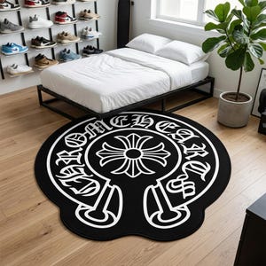 Könnte beinhalten: Ein runder schwarzer Teppich mit einem großen weißen Grafikdesign in der Mitte. Das Design zeigt ein stilisiertes Kreuzsymbol, das von einem kreisförmigen Rand mit dem Text CHROME HEARTS in einer fetten, gotischen Schriftart umgeben ist.