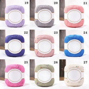 Puede incluir: Una colecci&oacute;n de nueve madejas de hilo en varios colores, incluyendo lavanda, gris, rosa, azul y verde. Cada madeja tiene una etiqueta blanca con un dise&ntilde;o ovalado. El hilo parece tener una textura moteada, con peque&ntilde;os adornos coloridos.