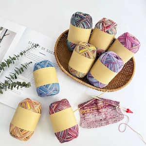 May include: A collection of colorful yarn skeins, each wrapped with a tan paper band, are arranged in a woven basket and on a white surface. A partially knitted piece with knitting needles is also present, suggesting a crafting theme.
