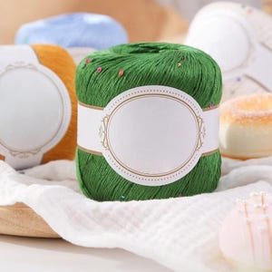 Puede incluir: Primer plano de ovillo de hilo verde con etiqueta blanca. El hilo es de color verde vibrante y tiene peque&ntilde;os adornos coloridos. Se ven otros ovillos de hilo en tonos naranja y azul en el fondo. El hilo est&aacute; sobre un pa&ntilde;o blanco.