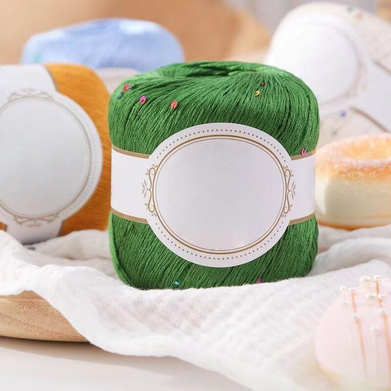 Puede incluir: Primer plano de ovillo de hilo verde con etiqueta blanca. El hilo es de color verde vibrante y tiene peque&ntilde;os adornos coloridos. Se ven otros ovillos de hilo en tonos naranja y azul en el fondo. El hilo est&aacute; sobre un pa&ntilde;o blanco.