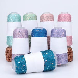 May include: A collection of colorful spools of thread, each wrapped with a band of sequins. The spools are in shades of teal, blue, purple, pink, and green. Each spool has a white label. The spools are arranged on a white surface.