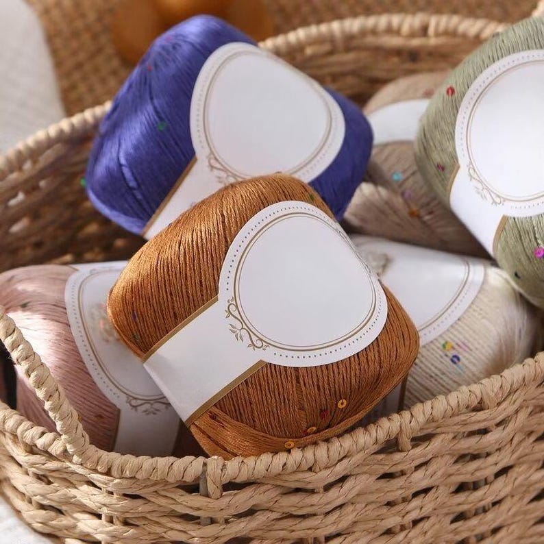 Puede incluir: Una cesta de mimbre llena de madejas de hilo de colores. El hilo es en tonos marr&oacute;n, azul, rosa y verde, cada uno envuelto con una etiqueta blanca. Algunas madejas tienen lentejuelas decorativas. La cesta est&aacute; hecha de material tejido natural.