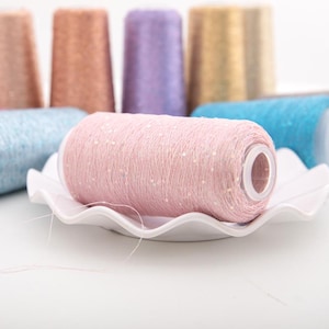 Può includere: Primo piano di una bobina di filo rosa con paillettes, appoggiata su un piatto bianco smerlato. Sullo sfondo sono visibili altre bobine di filo di vari colori, tra cui blu, oro e viola. Il filo è probabilmente per l'artigianato o il lavoro a maglia.