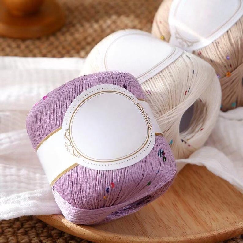 Puede incluir: Primer plano de tres madejas de hilo en tonos morado, crema y beige. La madeja morada est&aacute; en primer plano, con una etiqueta blanca. El hilo tiene motas de colores. Las madejas est&aacute;n sobre una bandeja de madera.