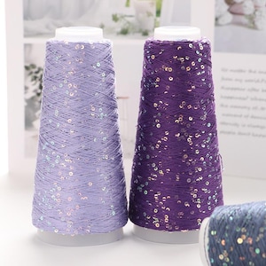 May include: Two cone-shaped spools of yarn, one lavender and one purple, are shown. Both are decorated with shimmering sequins. The yarn is wound around a white base. The background is softly blurred, focusing attention on the yarn's texture.