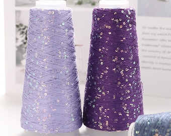 May include: Two spools of yarn, one lavender and one purple, are displayed. Both are cone-shaped and adorned with shimmering sequins. The yarn is wound around a white base, creating a visually appealing texture. The background is softly blurred.