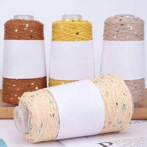 Puede incluir: Cuatro carretes de hilo en varios colores: marrón, amarillo, beige y beige claro. Cada carrete está adornado con lentejuelas. Los carretes tienen etiquetas blancas en el medio. El hilo está sobre una superficie blanca.