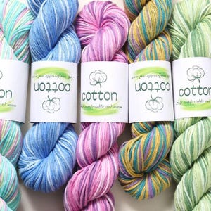 May include: Close-up of colorful cotton yarn skeins. The yarn is in shades of blue, pink, yellow, and green. Each skein has a white label with the word "cotton" and a cotton plant illustration. The text "Soft comfortable and warm" is also on the label.