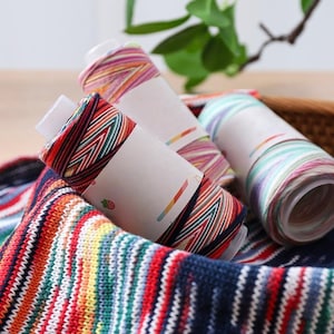 May include: Close-up of colorful yarn rolls with white labels, nestled in a woven basket. The yarn features a striped pattern in shades of red, blue, green, and yellow. The image showcases the texture and vibrant colors of the yarn.