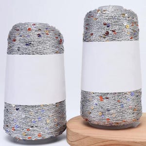 May include: Two spools of silver yarn with colorful sequins. Each spool has a white label and is set against a neutral background. The yarn is wound around a white core, creating a cone shape. One spool is on a wooden base.