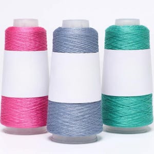 Könnte beinhalten: Drei Garnspulen in leuchtenden Farben: Pink, Blau-Grau und Türkis. Jede Spule hat ein weißes Etikett und einen transparenten Kunststoffdeckel. Das Garn ist eng gewickelt und erzeugt ein strukturiertes Aussehen. Ideal für Strick- und Häkelprojekte.