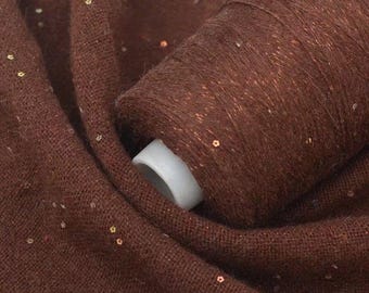 May include: Close-up of brown yarn on a white spool, with a brown fabric scattered around it. The yarn and fabric are speckled with small, shiny sequins. The yarn is tightly wound, and the fabric is soft and textured.
