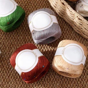 Puede incluir: Primer plano de varios ovillos de hilo en varios colores, incluyendo verde, gris, rojo y beige. Cada ovillo est&aacute; envuelto con una etiqueta blanca con un dise&ntilde;o ovalado. El hilo est&aacute; sobre una superficie marr&oacute;n texturizada, sugiriendo un tema artesanal o textil.