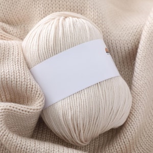 May include: A ball of cream-coloured yarn with a blank white label wrapped around it, set on a knitted fabric. The yarn is tightly wound, and the knitted fabric has a visible texture. The overall colour scheme is neutral, with an emphasis on texture and craft.